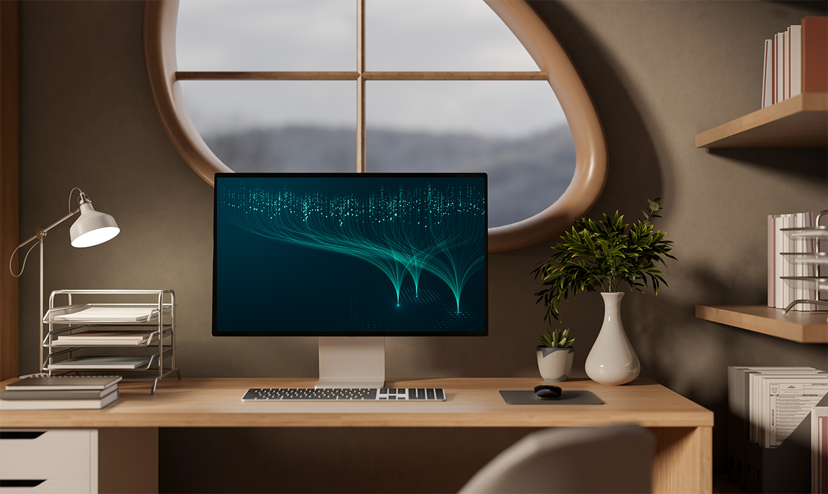 A computer monitor and keyboard on a tidy, modern wooden desk. A plant and bookshelf to the right, and lamp and papers and notebooks to the left.